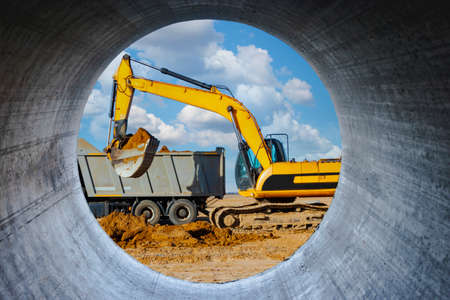 A powerful crawler excavator loads the earth into a dump truck against the blue sky. Development and removal of soil from the construction site. View through a large concrete pipeの写真素材