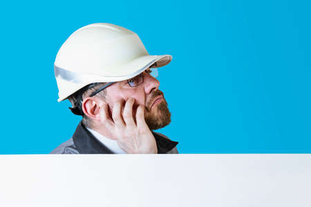 Positive friendly bearded male builder in overalls and safety hardhat and goggles looking at the...