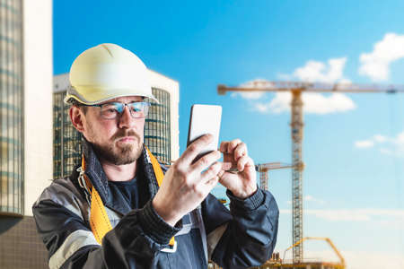 A male builder in a white hard hat against a blurred background of a construction site with a blue sky. Positive civil engineer with a beardの写真素材