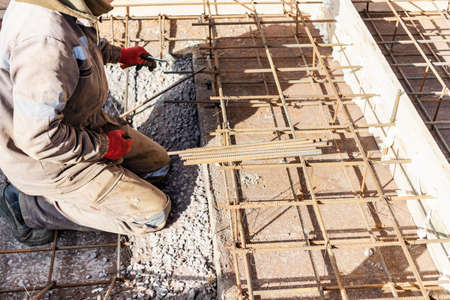 A worker uses steel tying wire to fasten steel rods to reinforcement bars. Reinforced concrete structures - knitting of a metal reinforcing cage. Preparation for concreting the foundationの写真素材