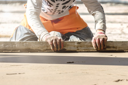 Close up of man builder placing screed rail on the floor covered with sand-cement mix at construction site. Male worker leveling surface with straight edge while screeding floor. Blurred backgroundの写真素材