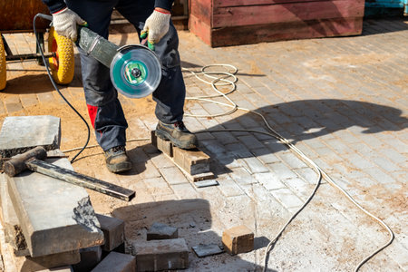 A construction worker using an electric grinder and a diamond cutting disc cuts paving slabs while...