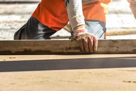 Close up of man builder placing screed rail on the floor covered with sand-cement mix at construction site. Male worker leveling surface with straight edge while screeding floor. Blurred backgroundの写真素材