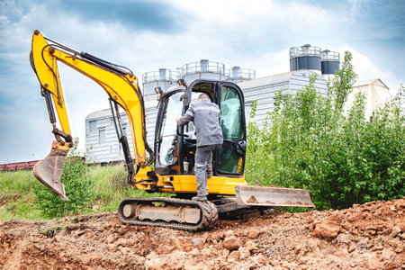 A man stands next to a excavator on a dirt field, surrounded by loose soil and construction equipment.の写真素材