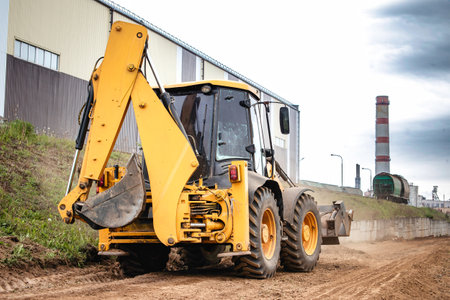 A large yellow construction vehicle or a loader is seen driving down a dusty dirt road, kicking up clouds of dust as it moves along.の写真素材