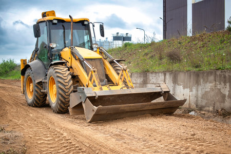 A tractor or a loader is parked on a dirt road, surrounded by fields and greenery under a clear sky.の写真素材