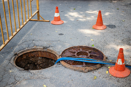 Workers are addressing a water supply failure by repairing an open sewer manhole surrounded by safety cones on a city street during the day.の写真素材