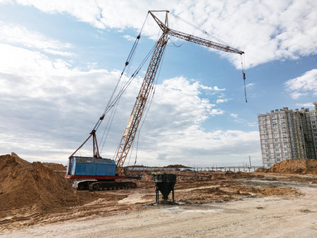 A crane lifts materials at a construction site amid earth mounds and a tall building in the background.の写真素材