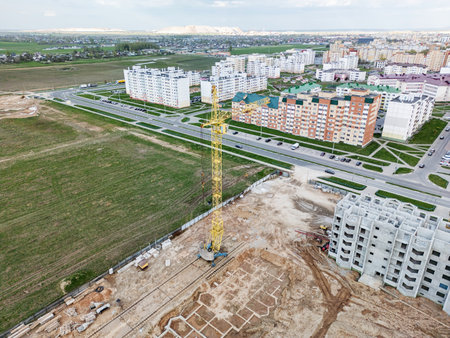 A yellow crane towers over a construction site, surrounded by residential buildings and lush green fields on a cloudy day.の写真素材