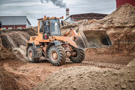 A construction vehicle digs earth on a cloudy day, highlighting activity at the site near a smoky factory.の写真素材