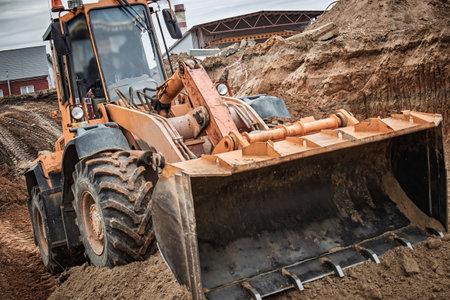 A large loader precision-digs in the soil, surrounded by dirt and construction materials on a cloudy day.の写真素材