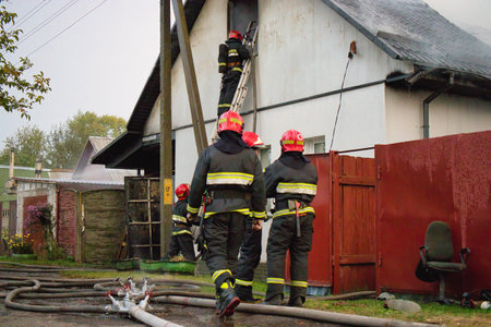 Firefighters work diligently to extinguish a fire, climbing ladders, managing hoses, and coordinating efforts to protect the nearby homes from danger.の写真素材