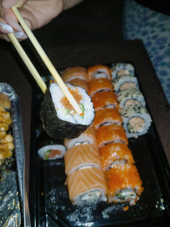 Chopsticks grasp a vibrant sushi roll filled with fresh ingredients, while an array of colorful maki awaits on a platter, perfect for sharing at dinner.の写真素材
