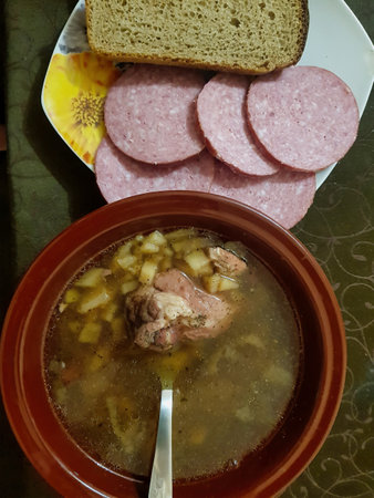 A steaming bowl filled with savory soup featuring tender meat and diced potatoes sits on a table. Accompanied by slices of cold cuts and fresh bread, this meal invites warmth.の写真素材