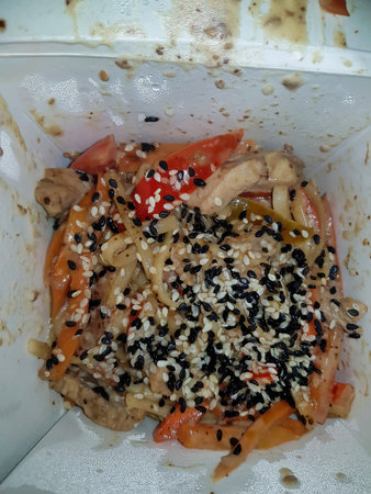 Savor a colorful dish of stir-fried chicken, vibrant vegetables, and a generous sprinkle of sesame seeds, enjoyed straight from a takeout container during a cozy evening.の写真素材