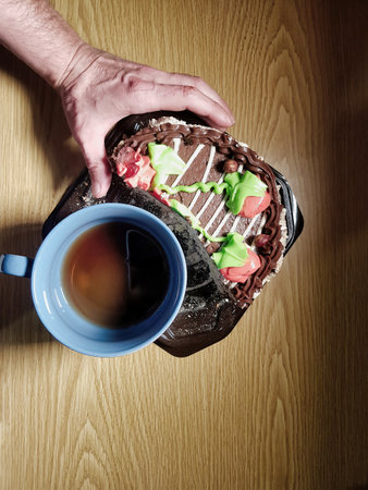 A hand holds a vibrant dessert adorned with frosting, accompanied by a steaming cup of tea on a rustic wooden surface, creating a cozy atmosphere.の写真素材
