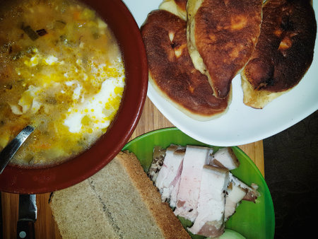 A hearty bowl of soup filled with vegetables and cream sits beside warm flatbreads and slices of cured meat, creating a comforting meal on a wooden surface.の写真素材