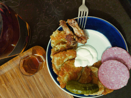 Crispy fried chicken is beautifully arranged on a plate with creamy sauce, slices of cured meat, dill pickles, and a small glass of amber liquor, setting a warm atmosphere.の写真素材