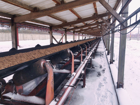 A long conveyor belt carries sand and materials through a covered structure as snow blankets the ground. A quiet winter day enhances the industrial atmosphere.の写真素材