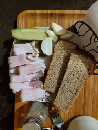 A rustic meal laid out on a wooden tray showcases thick slices of brown bread, succulent cured meat, crunchy pickles, and fresh onion wedges, evoking comfort and simplicity.の写真素材