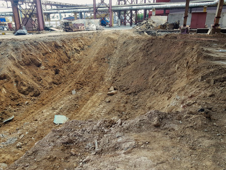 A large excavation area reveals freshly dug earth surrounded by machinery and debris, highlighting...