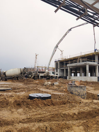 Heavy machinery operates at a construction site, pouring concrete for a large building. Workers coordinate amidst muddy terrain under a cloudy sky.の写真素材