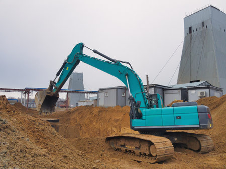 A powerful excavator digs into the soil at a bustling construction site. Silos rise behind, hinting at industrial activity in a gray, overcast sky.の写真素材