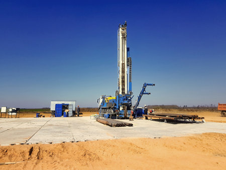 A drilling rig operates under the bright blue sky, surrounded by an arid landscape. Workers oversee the machinery as development projects take shape in this desolate area.の写真素材