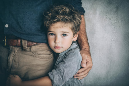 A young boy with a sad expression hugs his father's leg, seeking comfort and security against a soft, pastel backdrop, capturing a moment of connection.の素材
