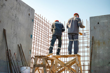 Two construction workers are positioned on wooden scaffolding, adding reinforcement to concrete walls under a clear blue sky.の写真素材