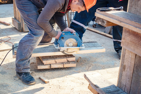 A worker in safety gear expertly uses a circular saw to cut wooden planks while surrounded by construction materials on a sunny day.の写真素材