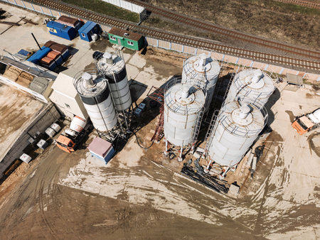Several large cement silos stand at a construction site adjacent to railway tracks, with machinery and containers scattered around.の写真素材