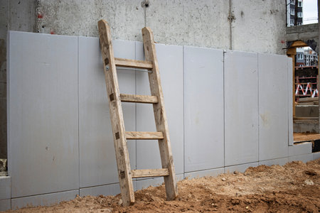 A wooden ladder leans against a gray panel wall at a construction site, with loose dirt surrounding it in an urban environment under bright daylight.の写真素材