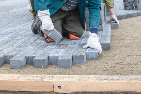 A worker kneels on the ground, carefully arranging gray paving stones in a pattern. Another person assists in the background, contributing to the project.の写真素材