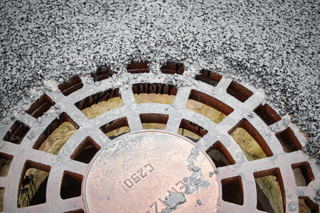 A rusty drainage grate is visible, embedded in fresh asphalt on a city road. The surrounding surface is smooth, showing recent roadwork taking place.の写真素材