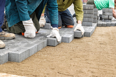 Workers are positioned on their knees, carefully arranging gray pavers on a sandy base during a construction project in clear weather.の写真素材