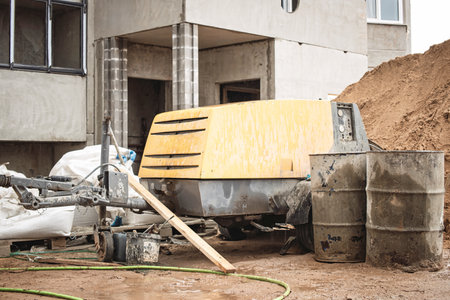 Heavy machinery is parked at a construction site where a structure is being built. Surrounding materials include barrels and stacked earth, indicating ongoing work.の写真素材