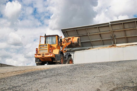 A construction vehicle maneuvers on a gravel surface, unloading materials at a work site, while...