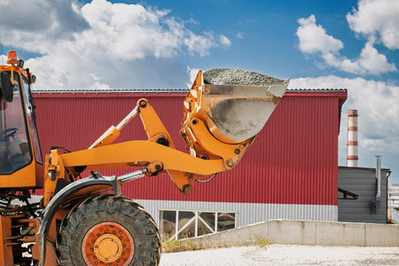 A heavy machine operates at an industrial location, lifting a bucket of gravel with a bright sky and...