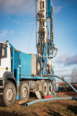 A large truck-mounted drilling rig is actively drilling in a rural landscape under a cloudy sky...