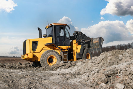 A loader or bulldozer moves large rocks on a job site. The sky is partly cloudy, indicating a...