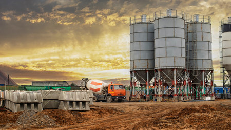A construction site features large cement silos and a mixer truck preparing for delivery of concrete...