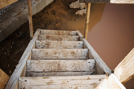 Sturdy but weathered wooden stairs descend into murky water at a construction site. The surroundings...