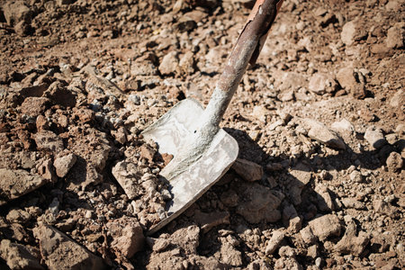 A metal shovel is placed on loose dirt at a construction site, indicating manual labor and...