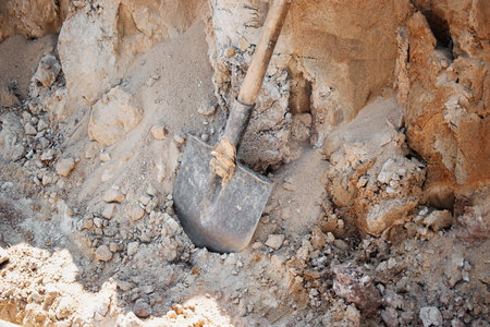 A metal shovel is embedded in loose dirt at a construction site, showing the ongoing earthwork in bright daylight. Surroundings reveal unfinished groundwork.の写真素材