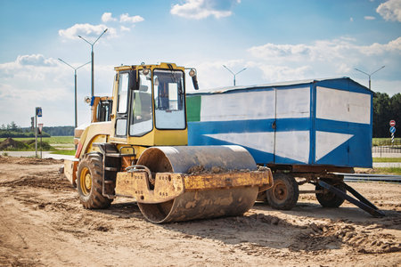 A large steamroller is working on a construction site, compacting freshly laid gravel under a clear...