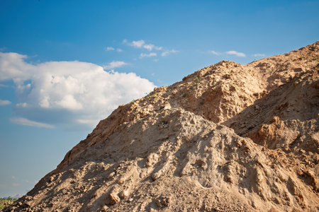 A large mound of dirt rises against a clear blue sky, showcasing earthy tones and textures with a few scattered clouds above, indicating a construction site in action.の写真素材