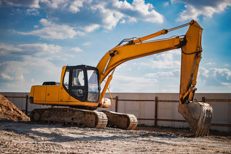 A yellow excavator stands on a construction site, ready for work. It is positioned on gravel, surrounded by earth mounds and a fence.の写真素材