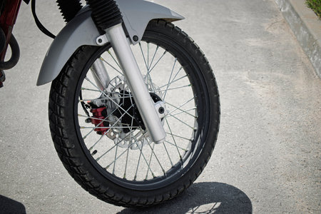 The close-up view shows a motorcycle wheel featuring spokes, a sturdy tire, and a disc brake, illuminated by sunlight on a concrete surface.の写真素材