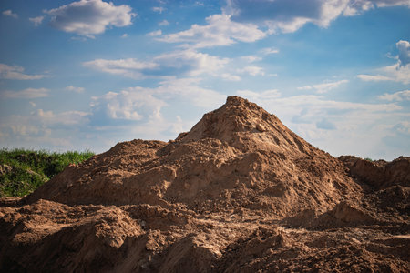 A large pile of dirt dominates the foreground, surrounded by a clear blue sky and scattered clouds. The setting reflects a construction site atmosphere.の写真素材